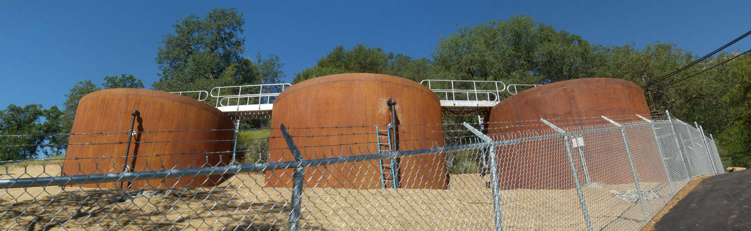 Three large rust-colored cylindrical storage tanks stand on a gravel slope, connected by a white catwalk; a barbed-wire chain-link fence separates them from a road beneath a clear blue sky.