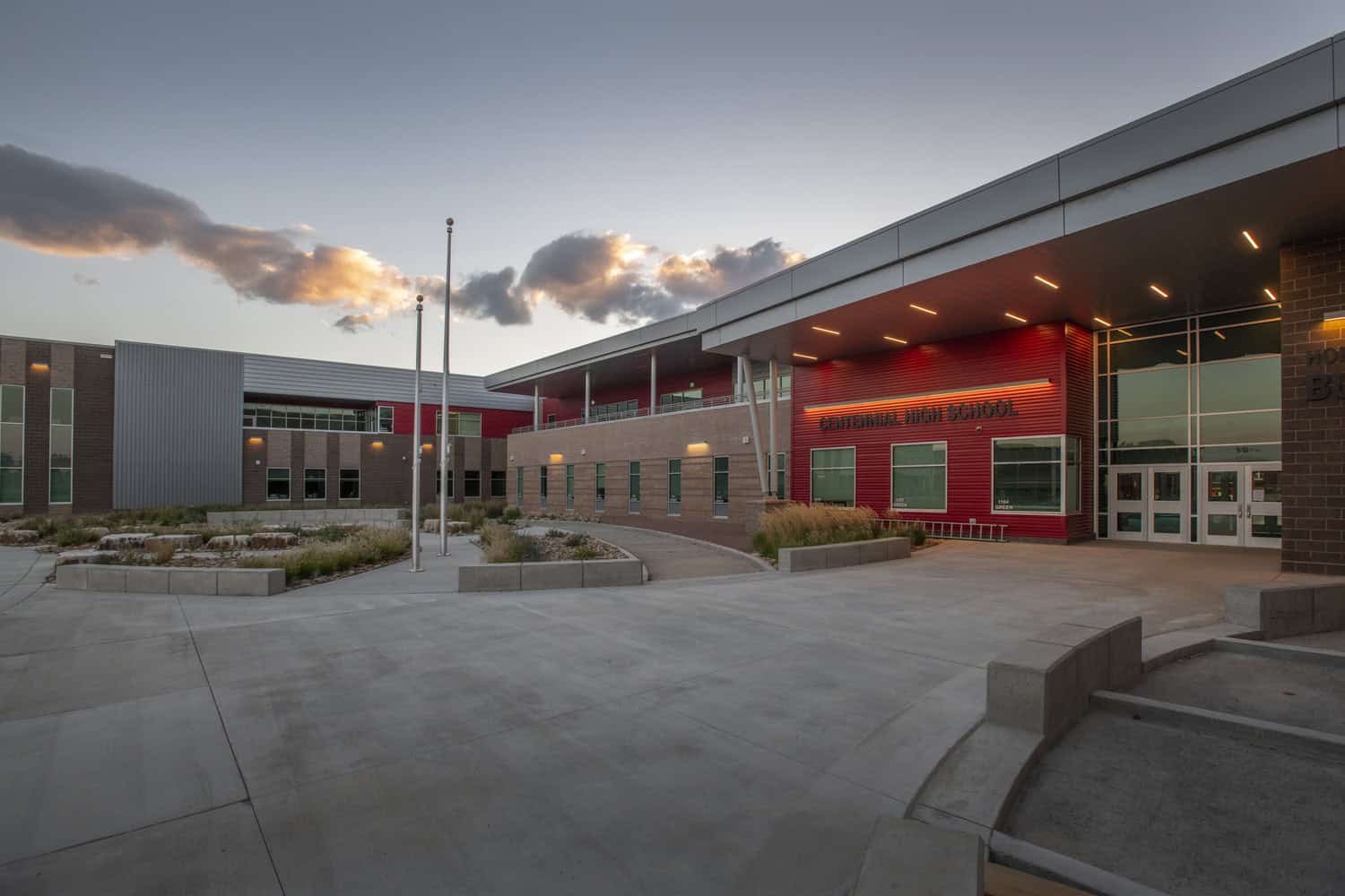 A modern red-and-brick high school building displays "CENTENNIAL HIGH SCHOOL" while exterior lights glow over an empty courtyard at dusk beneath scattered clouds.

Text found: "CENTENNIAL HIGH SCHOOL"