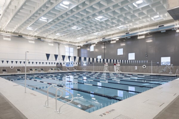 Indoor lap pool with calm, lane-separated water; starting blocks, lifeguard chairs, and triangular lane flags hang beneath a high, grid-patterned ceiling in a bright aquatic center.

Text visible: "3' 6"" NO DIVING (no diving symbol); "EXIT"