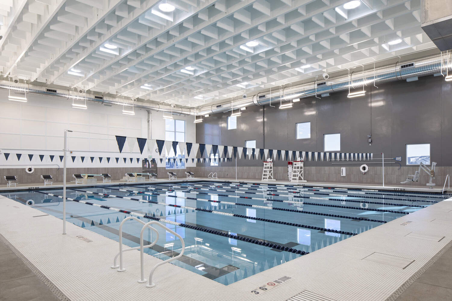 Indoor lap pool with calm, lane-separated water; starting blocks, lifeguard chairs, and triangular lane flags hang beneath a high, grid-patterned ceiling in a bright aquatic center.

Text visible: "3' 6"" NO DIVING (no diving symbol); "EXIT"