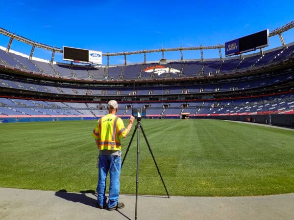 Worker in a fluorescent vest operates a tripod-mounted surveying scanner on the stadium turf; empty blue stands and a horse-logo of seats curve beneath a clear blue sky.

Text found: "Ford"; "EMPOWER FIELD"; "BUD LIGHT"; "FARO"