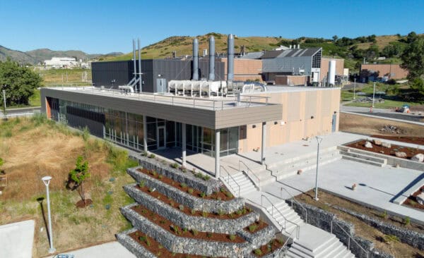 A low utility building sits atop terraced gabion stone planters and concrete steps, with rooftop ventilation stacks; surrounded by dry grass, paved plazas, adjacent industrial facilities and rolling green hills.