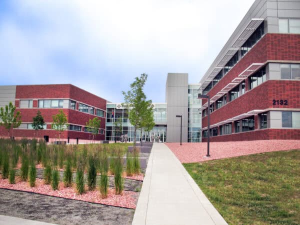 Brick office complex marked "2132" sits facing a central sidewalk, leading to a glass entrance; landscaping with grasses and young trees, lamp posts, and a cloudy sky overhead.