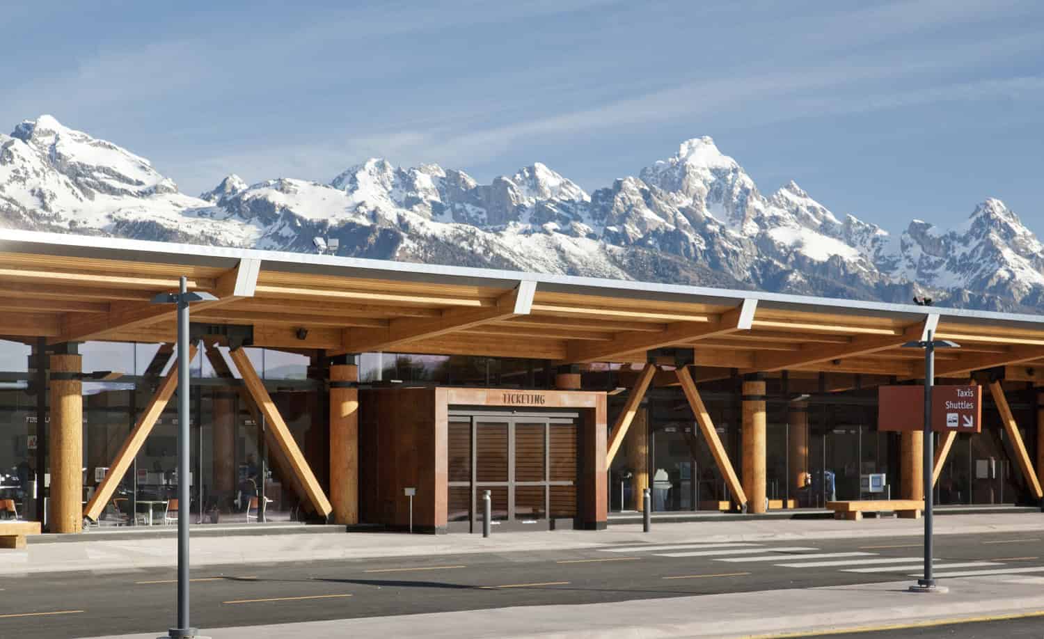 Wooden airport terminal houses closed "TICKETING" entrance, facing empty curbside; snow-capped mountains rise behind under a clear blue sky.

Text: TICKETING; Taxis Shuttles →
