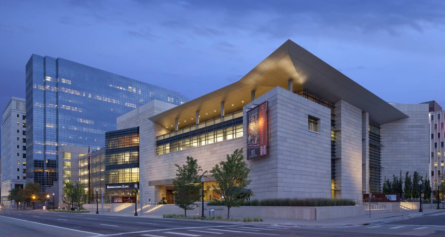 Modern limestone-and-glass cultural building sits lit at dusk, displaying a vertical banner and a glass-front café entrance, flanked by trees and a reflective glass office tower.

Visible text: "STORIES TOLD DAILY" ; "RENDEZVOUS CAFE" ; "NOW OPEN"