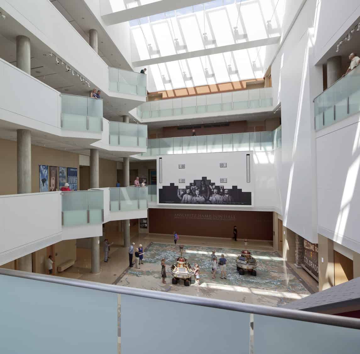 Multi-level atrium containing visitors who walk and view two small circular exhibits on a large floor map, beneath a skylight and glass railings, with a large black-and-white mural on the far wall.

Text: "ANSCHUTZ HAMILTON HALL"
"DEDICATED TO THE ENTREPRENEURIAL SPIRIT OF COLORADO"
"WELCOME TO COLORADO"
"LIVING WEST"