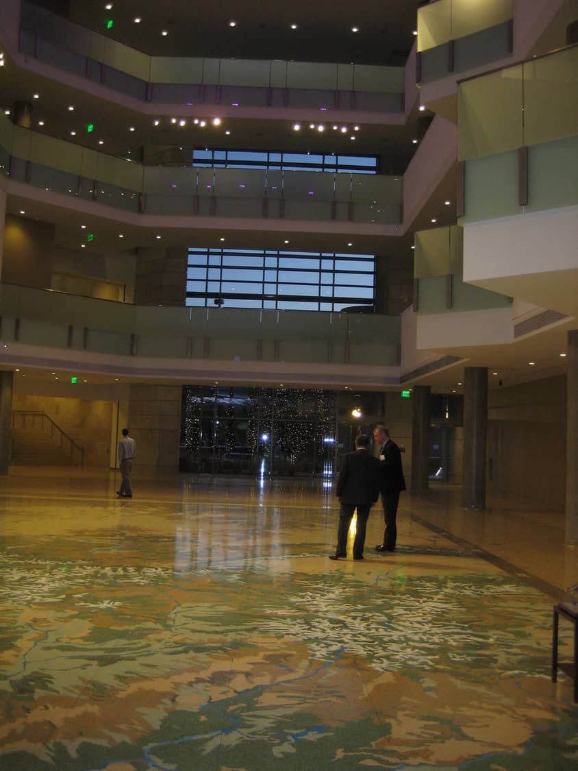 Two suited men converse on a large decorative floor map in a spacious multi-story atrium with glass balconies, overhead lights and a distant glass entrance; another person walks away.