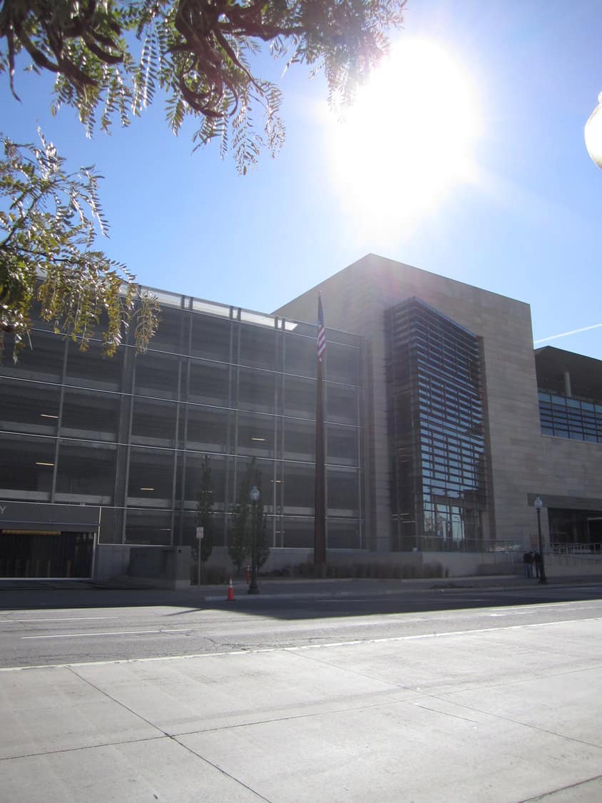 Multi-level parking garage and adjacent modern office building stand beside a city street as a bright midday sun shines overhead and an American flag hangs from a pole.