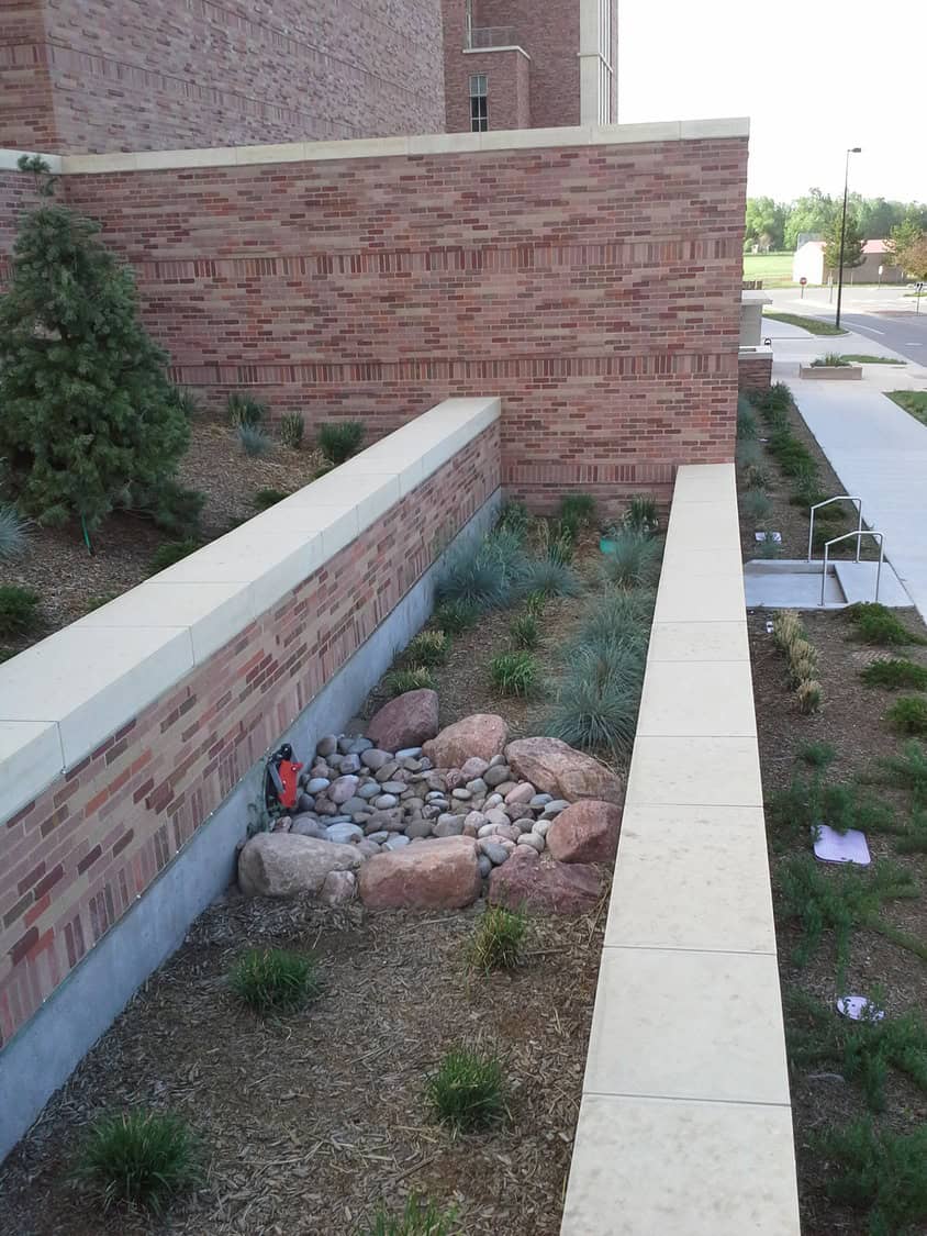 Rectangular brick planter contains a dry rock creek of boulders and river stones, with ornamental grasses and mulch, beside a brick building, sidewalk, and metal handrail.