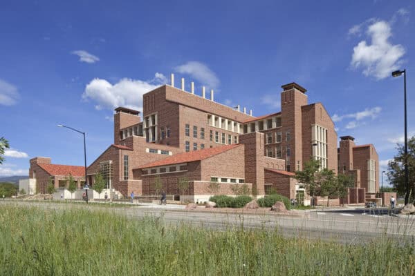 Red-brick multi-story academic building rises, featuring terracotta roofs, towers, and vertical windows; people walk and bike along the sidewalks in front, with grassy foreground and blue sky overhead.