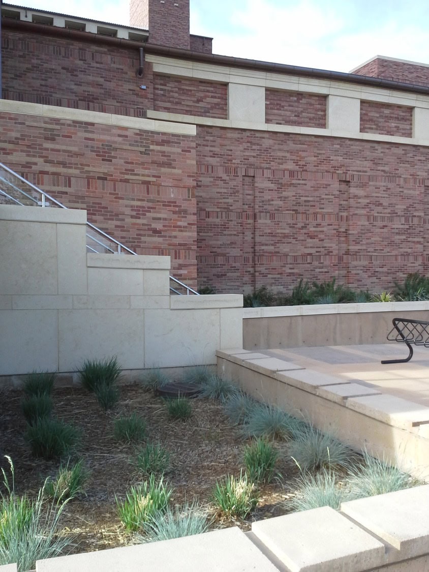 Low beige stone retaining walls and steps frame a planted bed of ornamental grasses; a metal bench sits on a raised paved platform against a tall red-brick building wall.