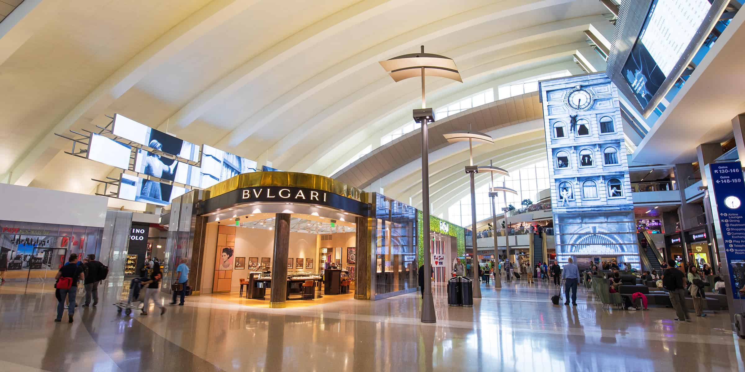 BVLGARI storefront gleams at center, open for browsing; travelers walk, wait and ride escalators beneath a vaulted airport concourse with large digital displays and lounge seating.

Text transcribed:
- BVLGARI
- Gates R 130-146  < 148-159
- Airline lounges
- Dining
- Restrooms
- Shopping
- Currency exchange
- Play areas
- Telephones
- Nursing room