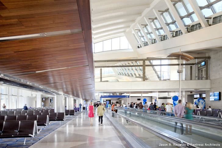 Airport terminal interior — passengers walk along and stand by a moving walkway amid rows of seating under a wood-paneled ceiling and high, sunlit clerestory windows.

Text in image: "Jason A. Knowles © Fentress Architects"