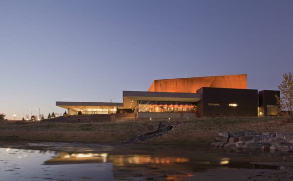 A contemporary angular building glows orange inside and out, its lights reflecting in a calm pond; set on a grassy shoreline with stone embankment under a clear twilight sky.
