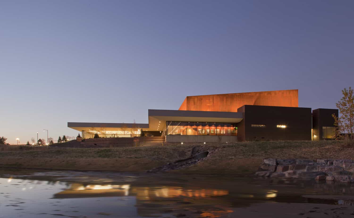 A contemporary angular building glows orange inside and out, its lights reflecting in a calm pond; set on a grassy shoreline with stone embankment under a clear twilight sky.