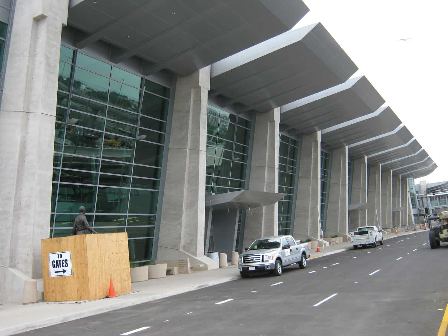 Modern airport terminal facade with tall concrete columns and overhanging metal canopy; pickup trucks parked along curb; a statue behind a plywood box; sign reads "TO GATES" →
