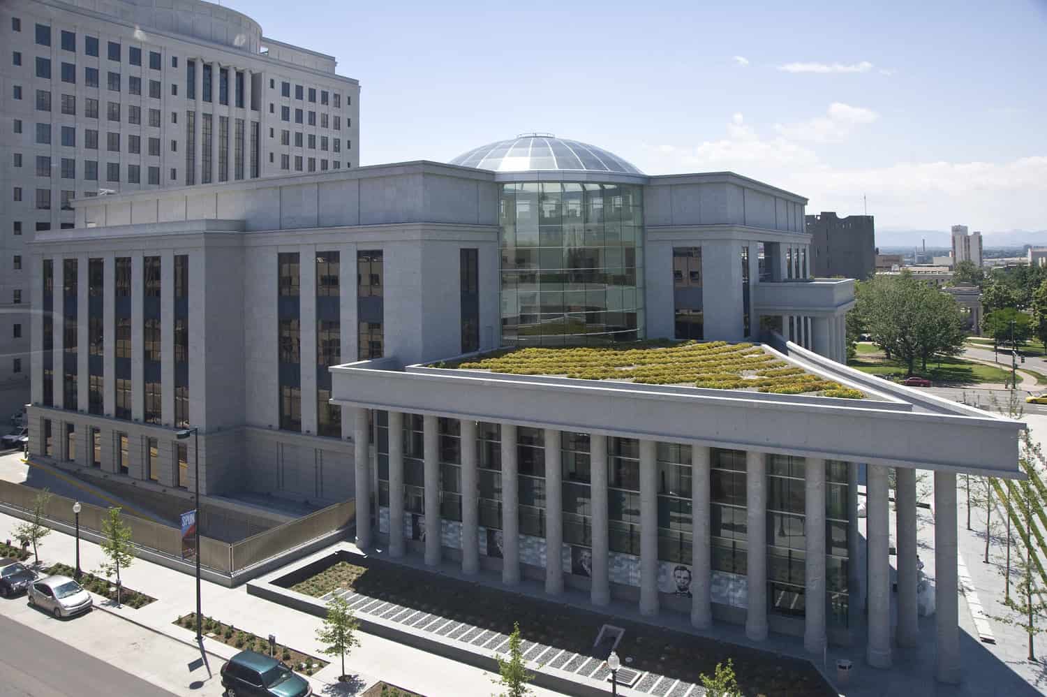 Large stone civic building features a glass-domed atrium and a planted triangular green roof, set on a downtown block with a colonnade, parked cars, and mature trees.