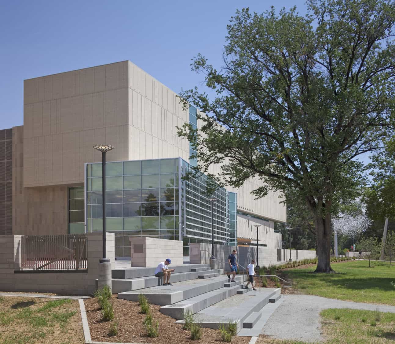 Modern beige-and-glass building rises behind tiered concrete steps where three people sit and walk, beside a large leafy tree and grassy lawn under a clear blue sky.