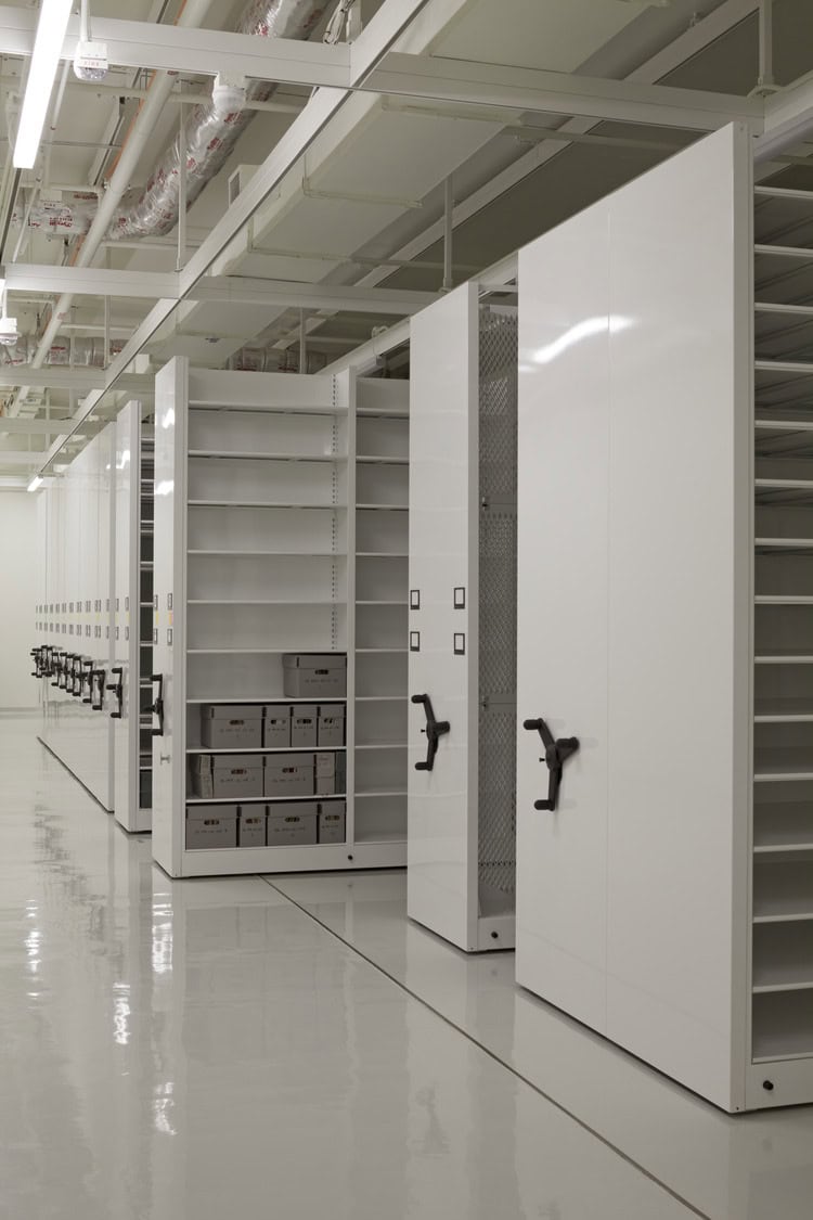 Mobile shelving units with hand cranks, some open to reveal empty shelves and stacked archive boxes, arranged along a polished floor under exposed ceiling ducts in a storage room.