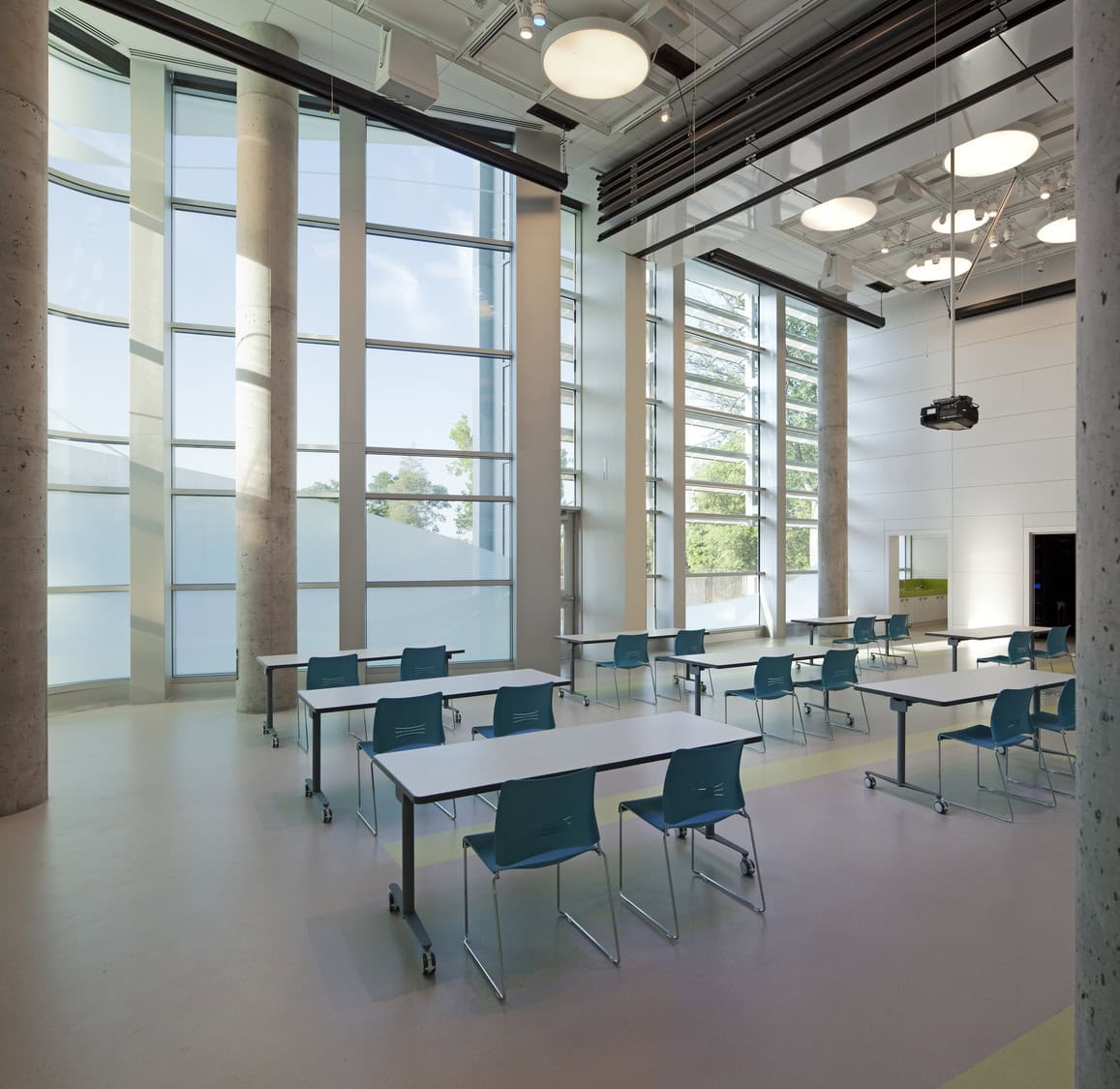 Rows of white tables with teal chairs sit spaced apart under hanging lights and a projector inside a high‑ceilinged modern classroom with tall floor‑to‑ceiling windows and concrete columns.