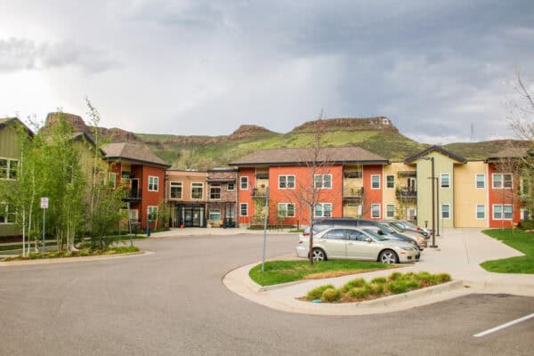 Apartment complex faces the camera; several cars are parked along a curved driveway; low green foothills and flat‑topped mesas rise behind under an overcast sky.