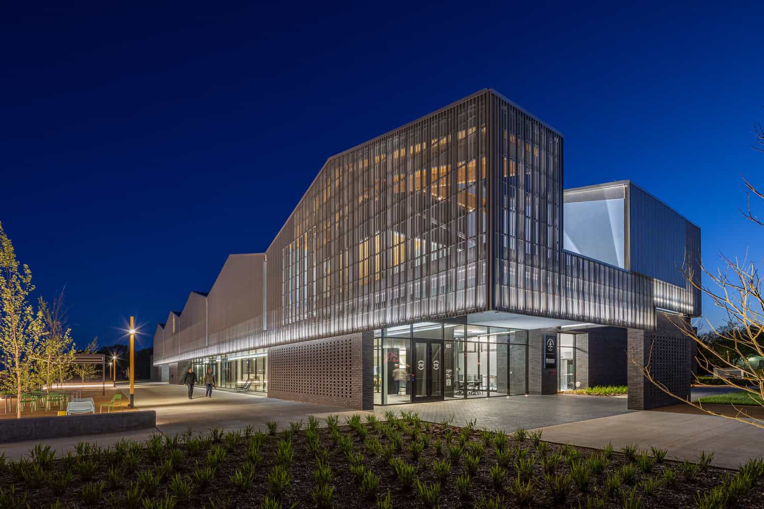 Contemporary glass-and-metal building glows as interior lights illuminate a perforated façade; two people walk past the entrance on a landscaped plaza at dusk. Text visible: "821".