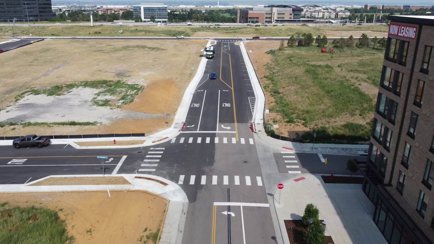 Road intersection (object) — a paved four‑lane street stretches away; action — a few cars drive and one is parked; context — vacant dirt and grassy lots flank it, mid‑rise building at right.

Text found: NOW LEASING; ONLY; ONLY; STOP