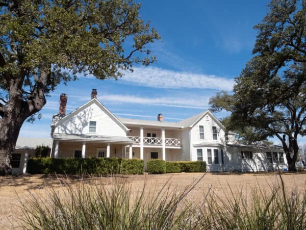 White two-story farmhouse sits on dry lawn, flanked by large oak trees; porch and balcony span the facade, chimneys rise from the roof under blue sky with wispy clouds.