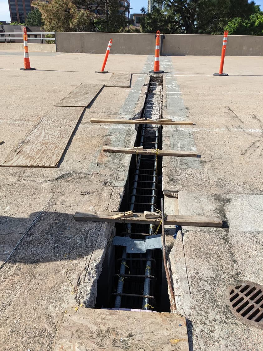 Long rectangular trench exposing rebar runs down a concrete parking-deck surface, supported by temporary wooden cross-braces and plywood; orange traffic cones block the area near a low parapet.