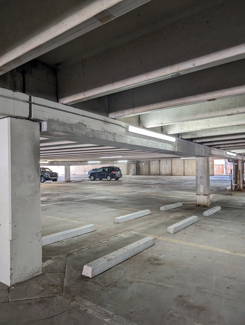 Concrete parking garage: empty parking bays with white wheel stops, two cars parked far back, exposed concrete beams overhead and fluorescent lights casting cool, dim illumination across stained floor.