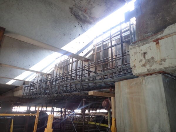 Rebar cage spans horizontally, being assembled for a concrete beam above scaffolding, with concrete slabs and columns surrounding the construction area and sunlight streaming through an overhead opening.