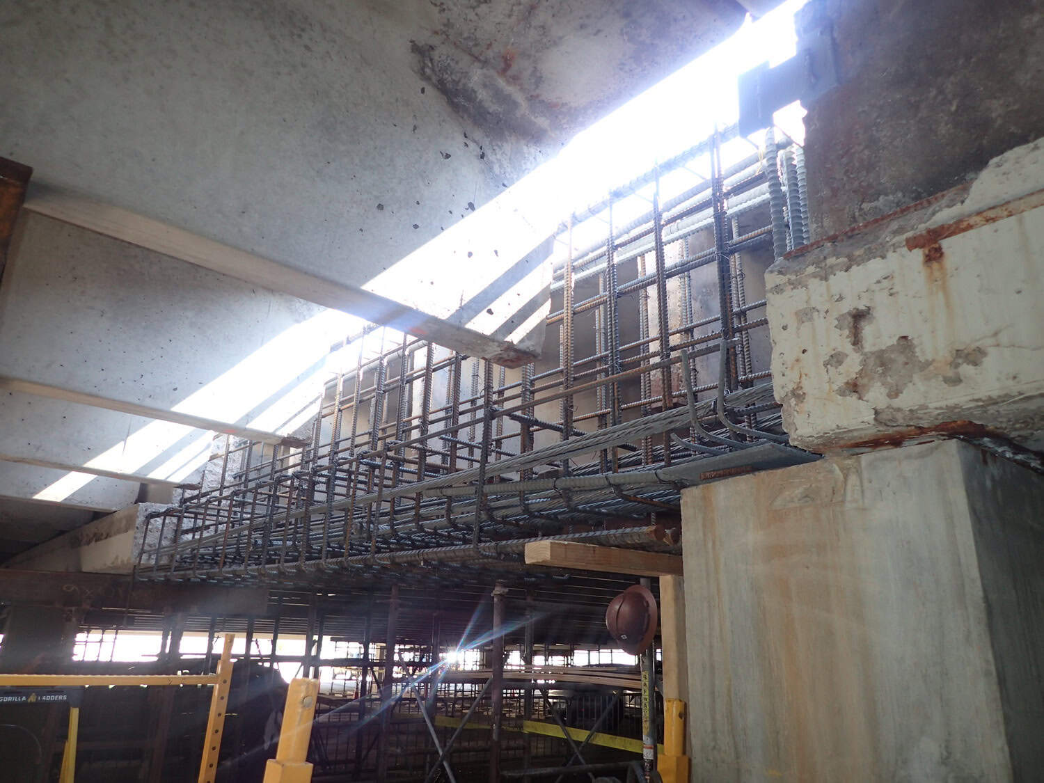 Rebar cage spans horizontally, being assembled for a concrete beam above scaffolding, with concrete slabs and columns surrounding the construction area and sunlight streaming through an overhead opening.