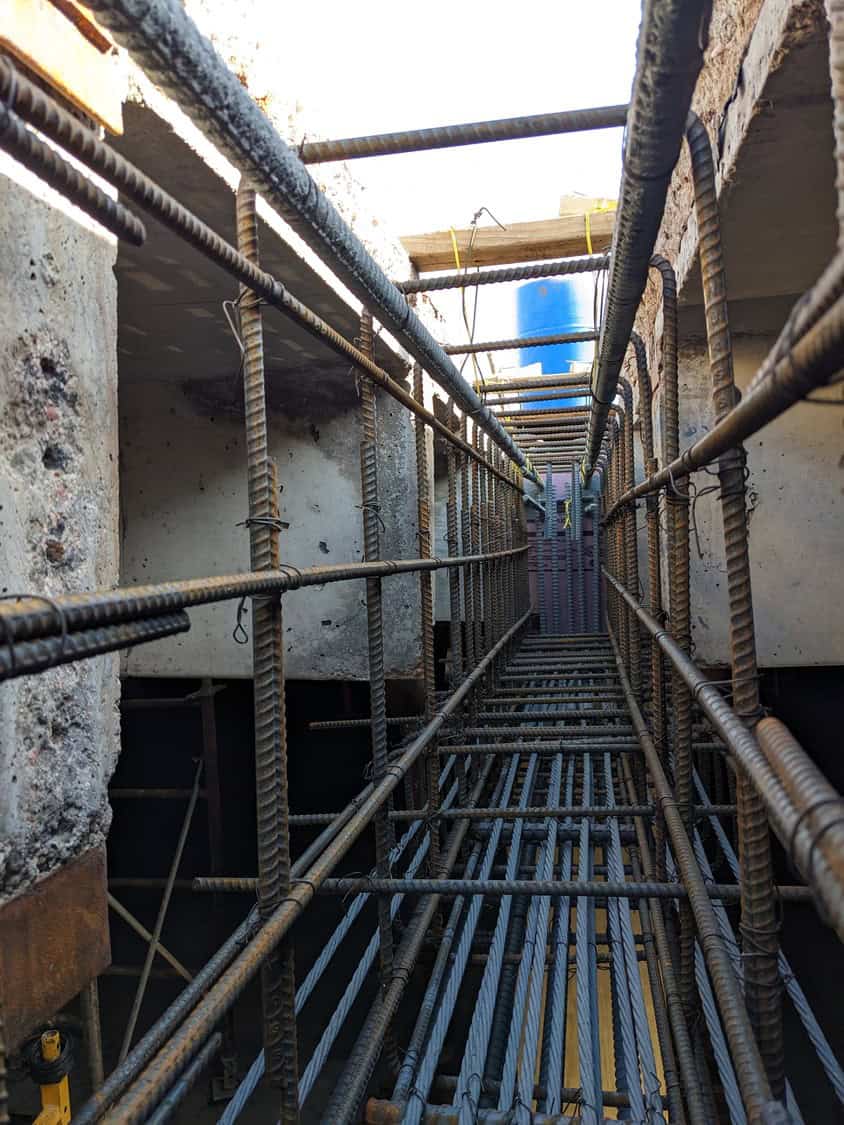 Rebar cage forming a narrow rectangular tunnel, reinforcing steel bars arrayed longitudinally and transversely, awaiting concrete pour inside a construction trench with concrete walls and a blue barrel above.