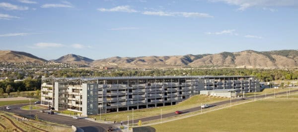 A multi-level parking garage sits on a grassy hillside; cars enter and exit on curved roads while pedestrians walk, framed by suburbs and dry rolling hills under a blue sky.