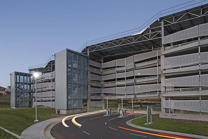 Multi-level parking garage with glass elevator towers; car light trails curve along an entrance road as vehicles enter at dusk, surrounded by grassy embankments and a clear sky.