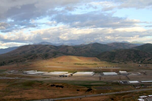 A sprawling industrial complex sits in a valley, illuminated by a patch of sunlight, surrounded by rolling brown-green hills and distant mountains under a broad cloudy sky.