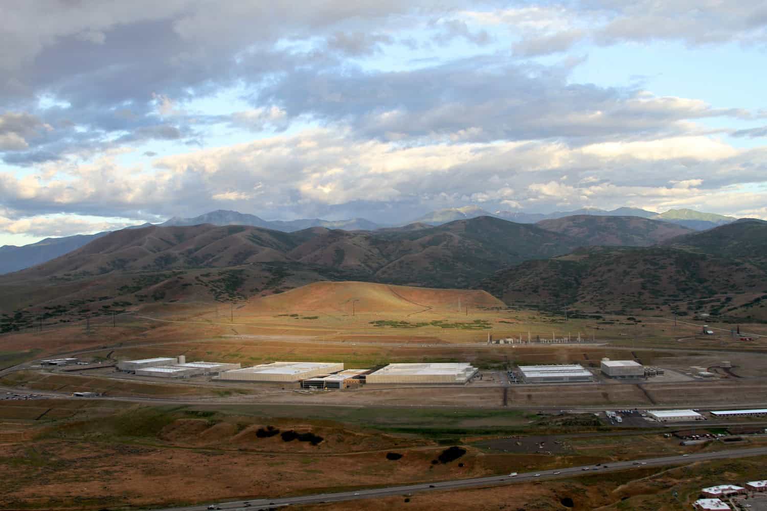 A sprawling industrial complex sits in a valley, illuminated by a patch of sunlight, surrounded by rolling brown-green hills and distant mountains under a broad cloudy sky.