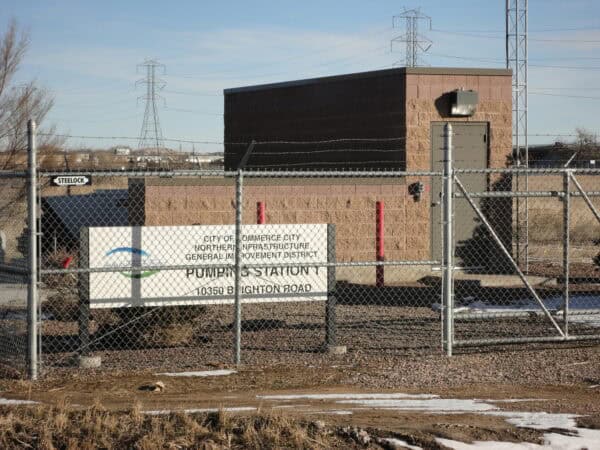 A chain-link fence with a posted sign encloses a small brick pump station building; gate, red bollards and a security light face a gravel lot with power towers and winter grass.

Transcribed text:
- STEELLOCK
- CITY OF COMMERCE CITY
  NORTHERN INFRASTRUCTURE
  GENERAL IMPROVEMENT DISTRICT
  PUMPING STATION 1
  10350 BRIGHTON ROAD
- (logo) WATER & SANITATION DISTRICT