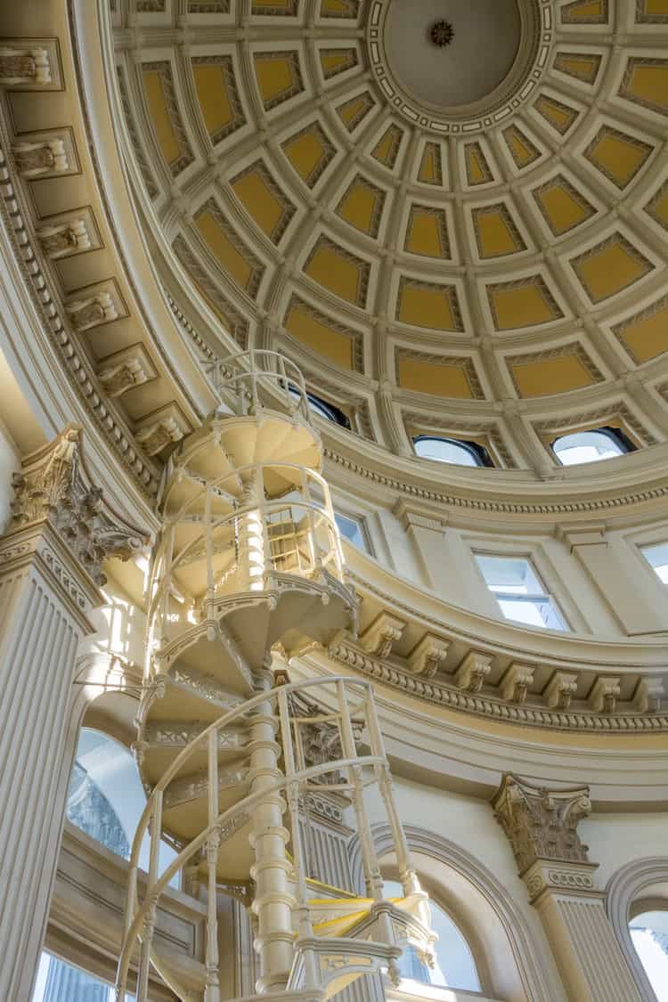 A cream-painted cast-iron spiral staircase (object) winds upward (action) toward a coffered dome inside a neoclassical rotunda with fluted columns, arched windows, and ornate cornices (context).