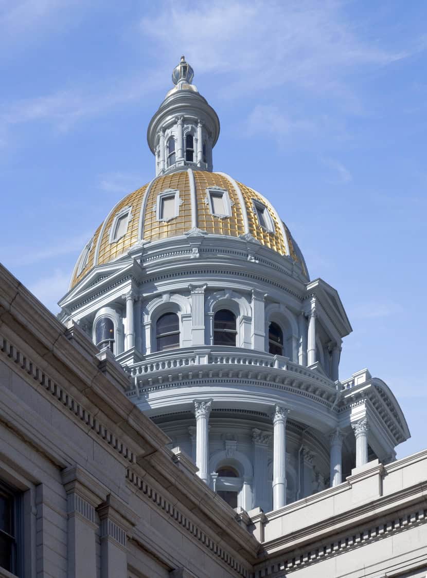 Gold-tiled dome crowns a pale-blue, colonnaded capitol drum, gleaming in sunlight above classical stone façades and cornices against a clear blue sky.