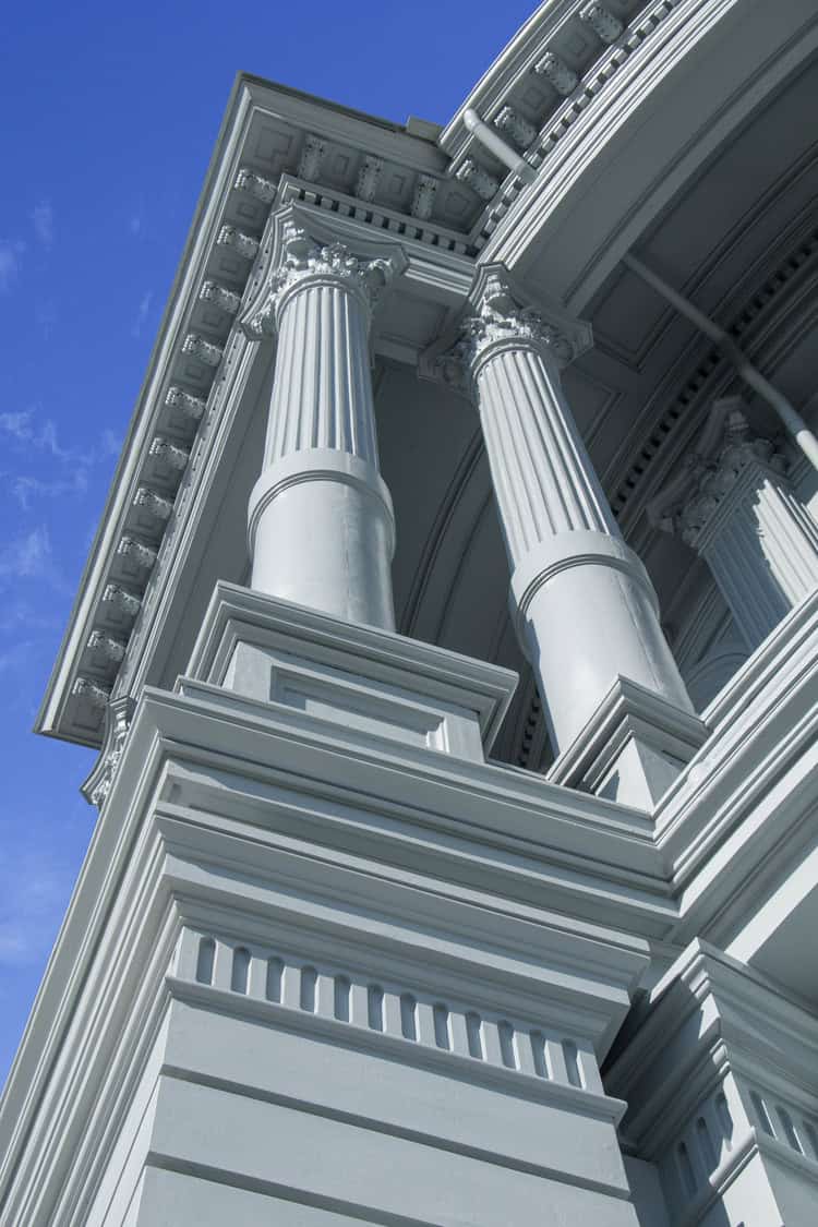 Neoclassical columns support an ornate entablature, photographed from below, emphasizing fluted shafts, Corinthian capitals and layered cornices against a clear blue sky.