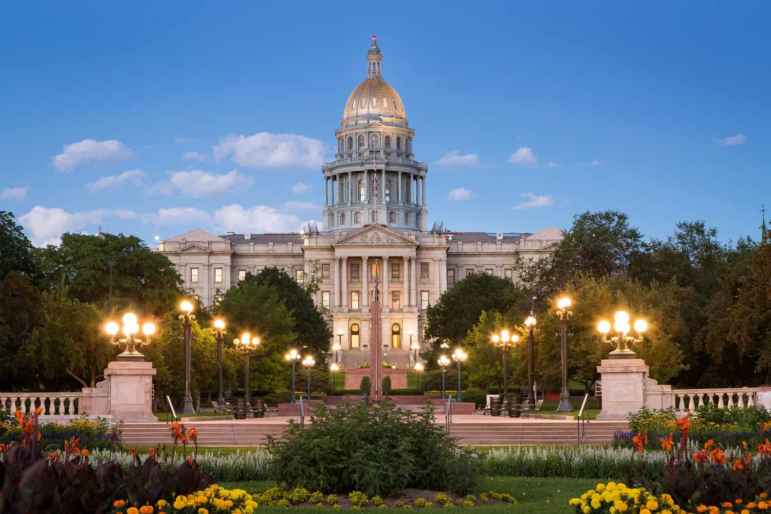 State capitol building with a gilded dome stands illuminated at dusk, flanked by lit lampposts, surrounded by manicured flowerbeds, trees, and a clear blue evening sky.