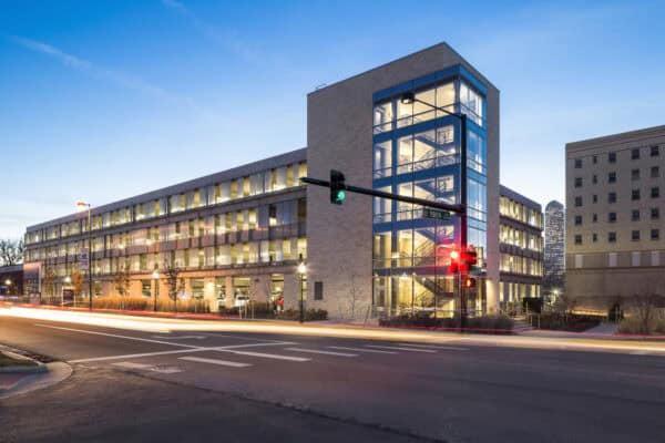 Glass-walled stair tower anchoring a lit multi-story parking garage, stairwell glowing while cars streak past; traffic lights show green and red at an urban intersection dusk — street sign reads "19th".