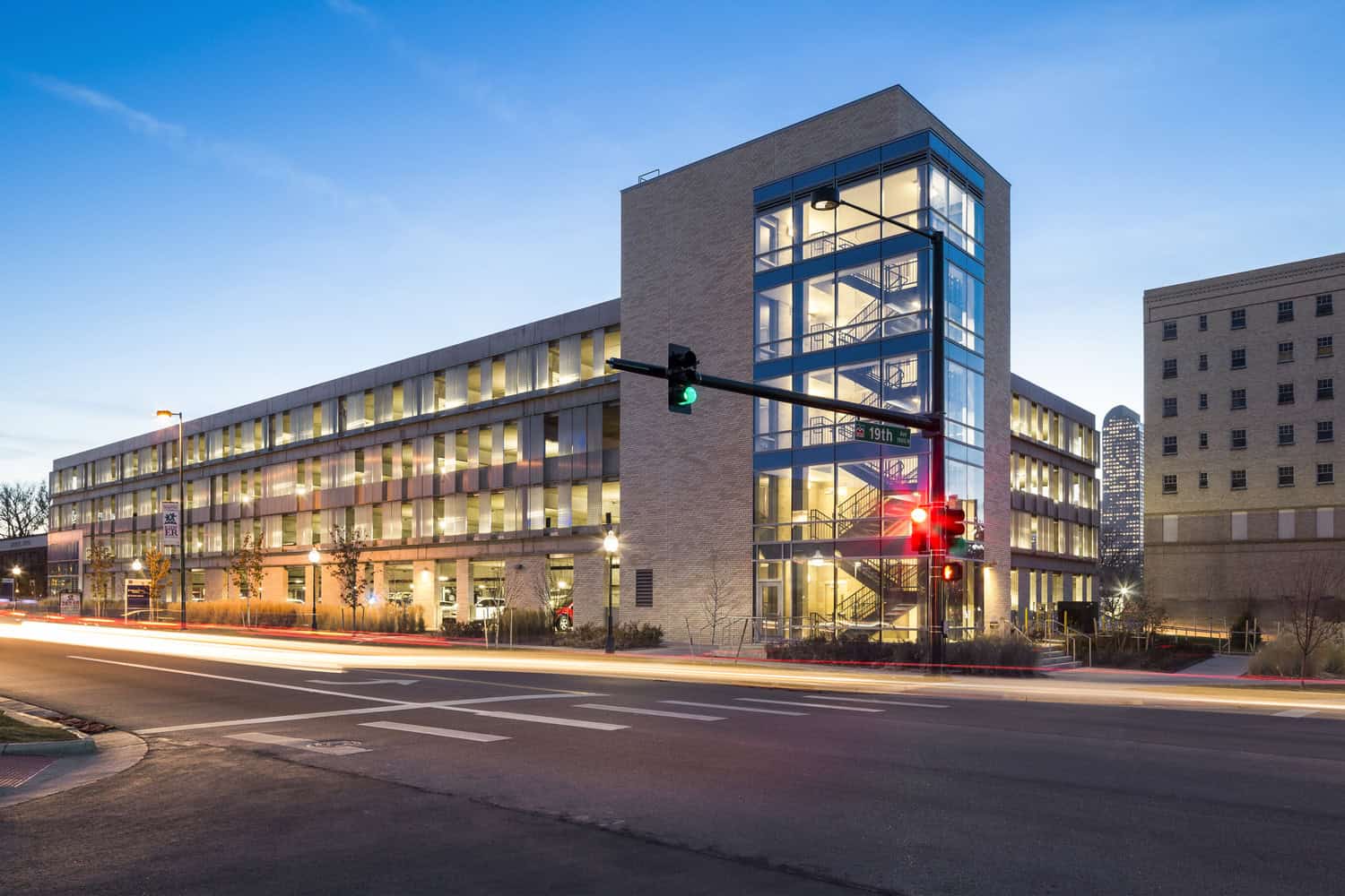 Glass-walled stair tower anchoring a lit multi-story parking garage, stairwell glowing while cars streak past; traffic lights show green and red at an urban intersection dusk — street sign reads "19th".