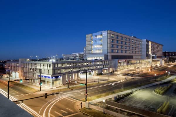 Hospital building illuminated from within, emergency entrance lit; vehicle light trails curve through a busy urban intersection at dusk beside a multi-level parking structure.  
Text visible: "EMERGENCY", "Downing St"