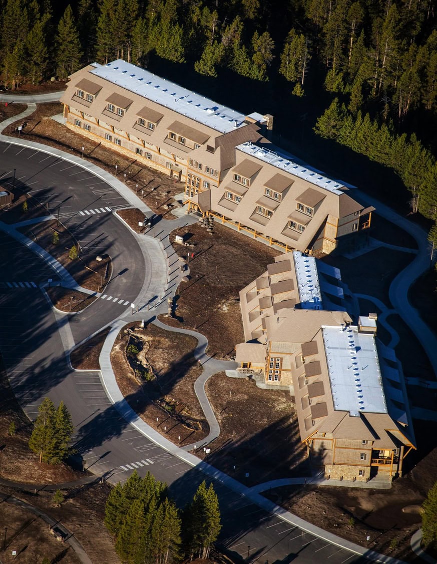 Three long, multi-story lodge buildings line a curved driveway and empty parking area, connected by walkways and courtyards, set in a cleared lot bordered by dense pine forest.