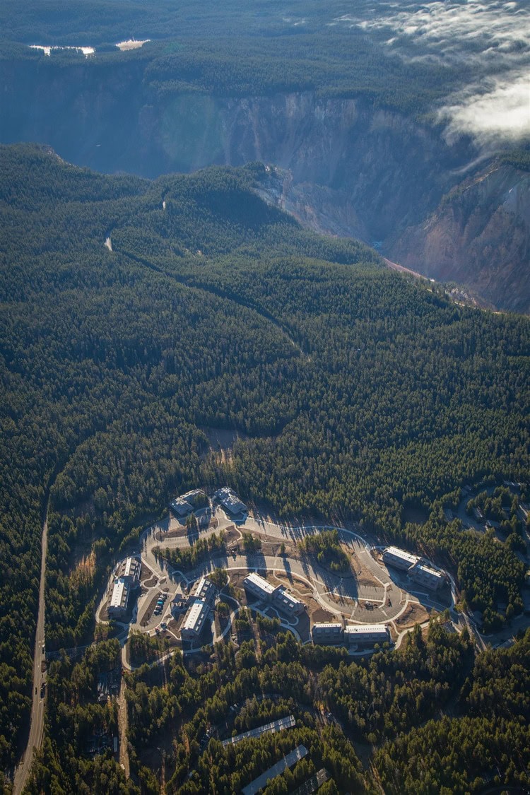 A cluster of low-rise buildings sits in a clearing, connected by curving roads, surrounded by dense pine forest at the edge of a steep canyon beneath scattered clouds.