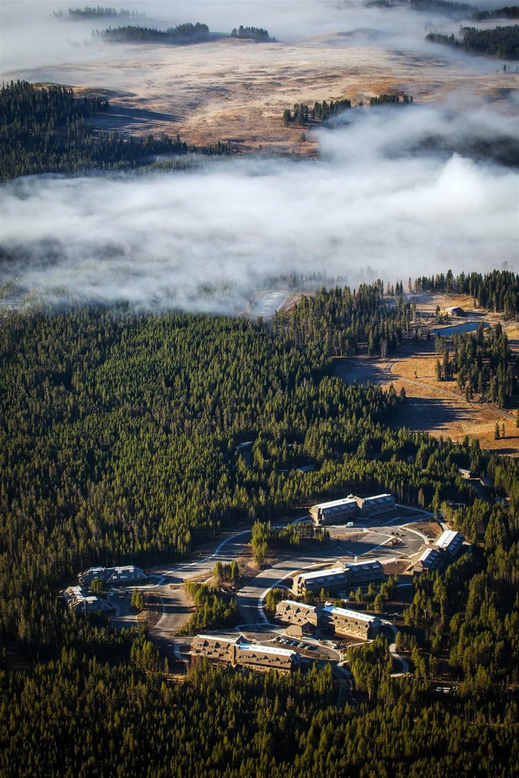 A cluster of lodge-like buildings sits nestled in dense conifer forest; low fog drifts across adjacent open meadows and valleys under soft morning light in a mountainous setting.