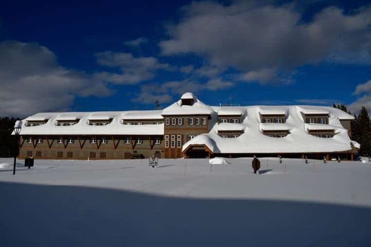 A large multi-story wooden lodge sits blanketed under deep snow, with drifts on roofs; a lone person and lamppost stand in a snowfield, evergreens and blue sky behind.
