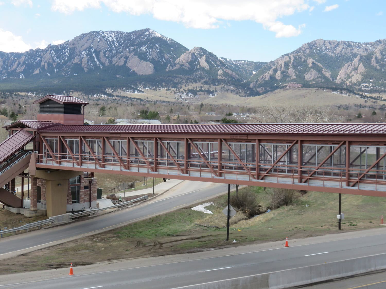 Covered pedestrian bridge spans a road, connecting a stair tower to a sidewalk; mountains with patches of snow rise in the background, with grassy roadside and traffic cones below.